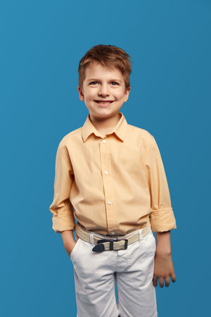 Vertical photo of handsome cute child boy wearing trendy beige shirt, smiling for camera while keeping hand in pocket against blue background.Children studio portrait. Childhood lifestyle conceptの写真素材