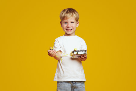 Charming little blonde boy smiling happily while holding a camera photo, standing isolated over yellow background.の写真素材