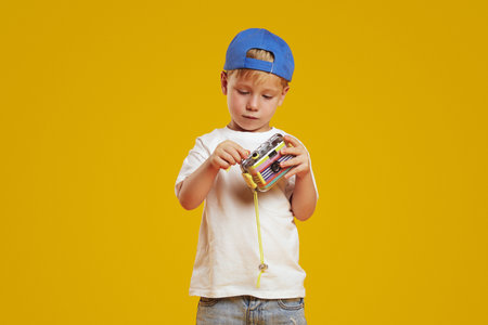 Adorable little boy wearing white shirt and blue cap holding photo camera with curiosity while standing against yellow background.の写真素材