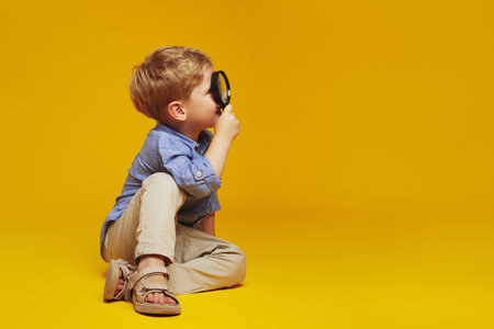 Horizontal photo of cheerful boy with blonde hair sitting on studio floor while looking through magnifier at free empty space for text, blank. Isolated over yellow background.の写真素材