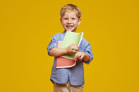 Excited little nerd kid in blue shirt smiling and holding two notebooks while being ready for...