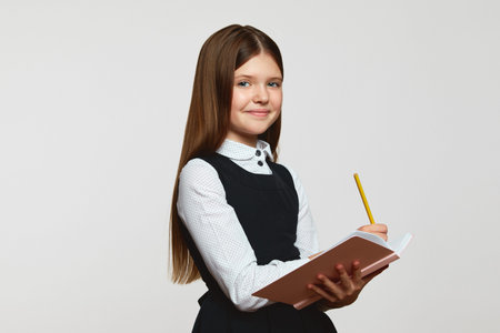 Cheerful elementary schoolgirl in uniform holding open notebook and looking at camera, standing...