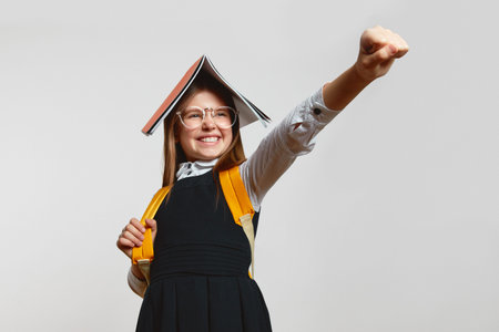 Funny girl in school uniform and eyeglasses holding book on head while acting like superhero with clenched fist outstretched on white backgroundの写真素材