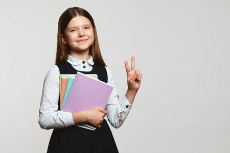 Clever little nerd girl with textbooks in school uniform showing v sign and looking at camera...