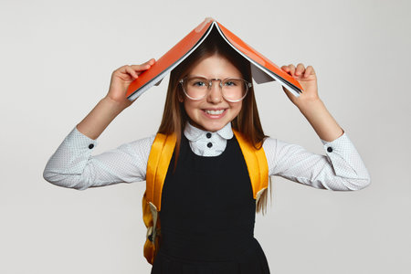 Positive little blonde girl wearing eyeglasses and yellow backpack holding open copybook on head while smiling broadly against white backgroundの写真素材