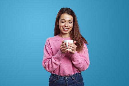 Dreamy beautiful young Hispanic woman holds cup of coffee, drinks hot beverage and imagines pleasant moment wears pink jumper, isolated against blue backgroundの写真素材