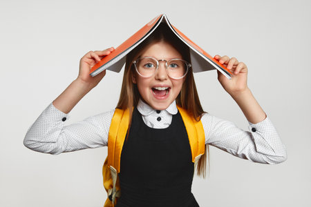Cheerful child in school uniform and yellow backpack covering head with book during lesson against white backgroundの写真素材