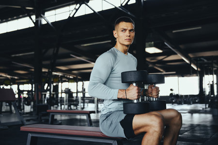 Strong fit man sitting on gym bench holding dumbbells, ready for strength training. Focused and...