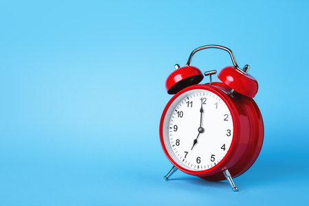 A red retro alarm clock stands against a vibrant blue background, symbolizing time management and punctuality. The clock is set at 12 oclock, evoking a sense of urgency and attention.の写真素材