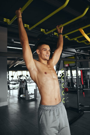 Vertical photo of muscular strong shirtless man doing abs workout on bar in modern gym