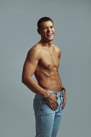 Fit and cheerful shirtless male showcasing a healthy and athletic physique, standing confidently while smiling against grey background. Demonstrates happiness, health, and an active lifestyle.の写真素材