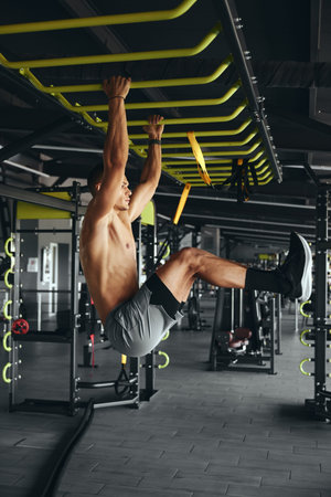 Vertical photo of muscular strong shirtless man doing abs workout on bar in modern gym