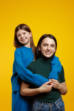 Portrait of two cheerful sisters hugging and smile looking at camera, isolated over yellow backgroundの写真素材