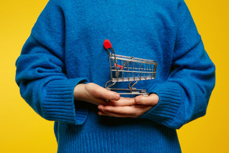 Unrecognizable young child wearing a bright blue sweater carefully holds a tiny shopping cart, standing against a vivid yellow backdrop. Ideal for retail advertising and children marketing campaigns.の写真素材