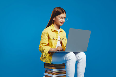 Attractive little young kid girl intently browsing on a laptop, seated atop a stack of books,...