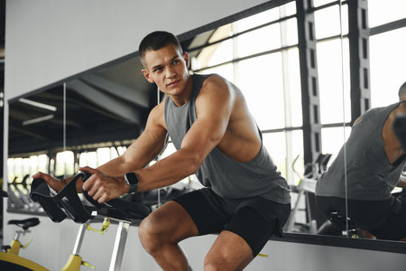 Athletic guy cardio riding on bicycle at gym fitness and looking aside. Muscular people workout on...