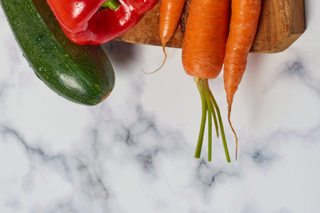 Various greens and vegetables on wooden kitchen table a white marble surface. Carrots, peppers,...