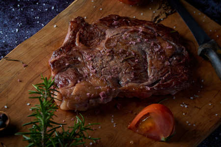 Grilled beef steak on wooden board with salt and other seasonings on a dark surface. Steak ready for eating. Dark food. Meat, tomato, pepper, lemon and rosemary.の写真素材