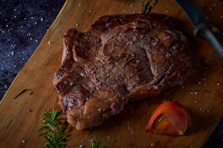 Grilled beef steak on wooden board with salt and other seasonings on a dark surface. Steak ready for eating. Dark food. Meat, tomato, pepper, lemon and rosemary.の写真素材
