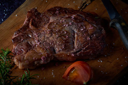 Grilled beef steak on wooden board with salt and other seasonings on a dark surface. Steak ready for eating. Dark food. Meat, tomato, pepper, lemon and rosemary.の写真素材