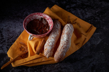 Fartons, typical pastries from Valencia, Spain, with hot chocolate, in a Chinese ceramic container on top of a yellow cloth, with a dark surface. A sweet breakfast or snack.の写真素材
