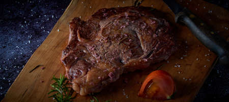 Grilled beef steak on wooden board with salt and other seasonings on a dark surface. Steak ready for eating. Dark food. Meat, tomato, pepper, lemon and rosemary.の写真素材