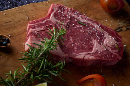 Fresh and raw beef steak on wooden board with salt and other seasonings on dark surface. Steak ready to grill. Dark food. Meat, tomato, pepper, lemon and rosemary.の写真素材