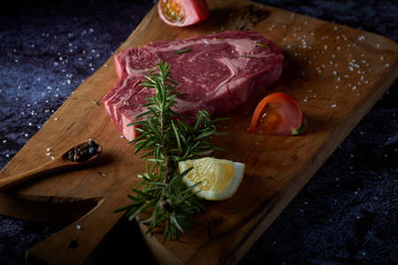 Fresh and raw beef steak on wooden board with salt and other seasonings on dark surface. Steak ready to grill. Dark food. Meat, tomato, pepper, lemon and rosemary.の写真素材