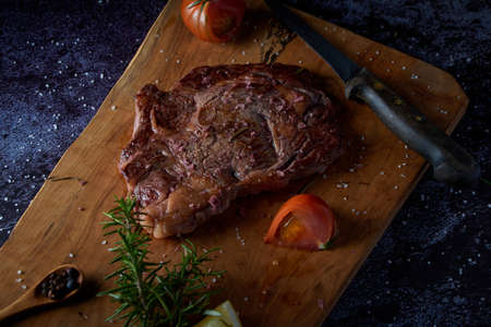 Grilled beef steak on wooden board with salt and other seasonings on a dark surface. Steak ready for eating. Dark food. Meat, tomato, pepper, lemon and rosemary.の写真素材