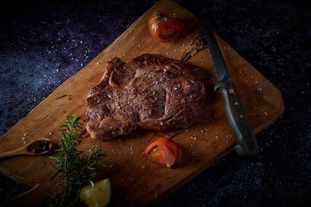 Grilled beef steak on wooden board with salt and other seasonings on a dark surface. Steak ready for eating. Dark food. Meat, tomato, pepper, lemon and rosemary.の写真素材