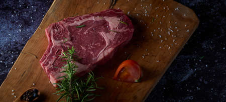Fresh and raw beef steak on wooden board with salt and other seasonings on dark surface. Steak ready to grill. Dark food. Meat, tomato, pepper, lemon and rosemary.の写真素材