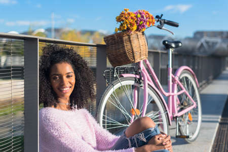 Black happy young woman sitting by the river with her pink vintage bicycle with basket flowers  の写真素材
