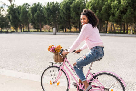 Black happy young woman riding a vintage bicycle with beautiful basket of flowers.の写真素材