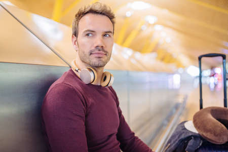 Young man waiting listening music and using mobile phone at the airport with a suitcaseの写真素材