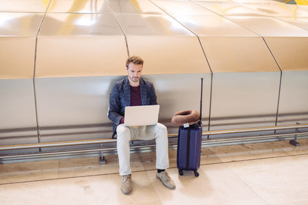 Young attractive man sitting at the airport working in his laptop waiting his flight with a suitcase.の写真素材
