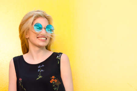 happy young woman smiling on a yellow wall with sunglassesの写真素材