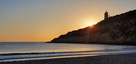 Deserted beach one summer day when the day goes and the sunset produces a backlight on a mountain with a lighthouse at the top.の写真素材