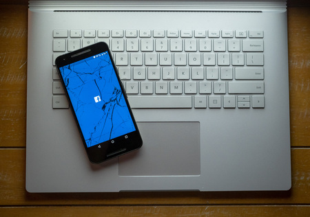 APRIL 8, 2018: A cracked phone with the Facebook app loading sitting on a laptop. The social media giant has been in hot water for privacy troubles.のeditorial素材