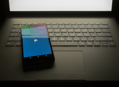 APRIL 8, 2018: Phone sitting on laptop with Facebook desktop site reflecting on screen. The social media giant is in govenrment trouble for the Cambridge Analytica scandal.のeditorial素材