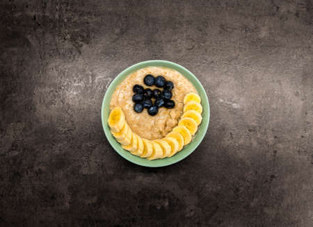Bowl of oatmeal oatmeal with banana and blueberry on vintage table in flat gray weighted style. Healthy breakfast and diet food.の写真素材