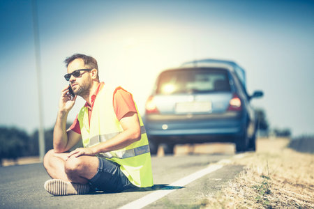 Man sitting on the road talking by phone after a car breakdown.の写真素材