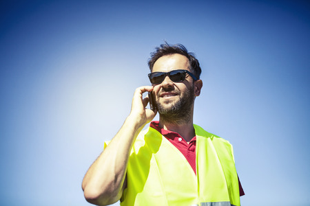 Engineer with reflective vest and sunglasses talking by phone. Blue sky background.の写真素材