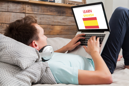 Young man using a laptop for learning spanish while lying in the bed at home.の写真素材
