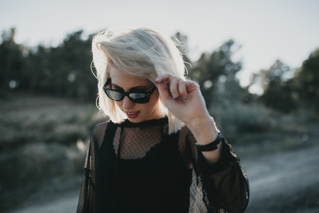 Close up portrait of blonde woman with sunglasses and black dress walking outdoors with sun backlighting.の写真素材