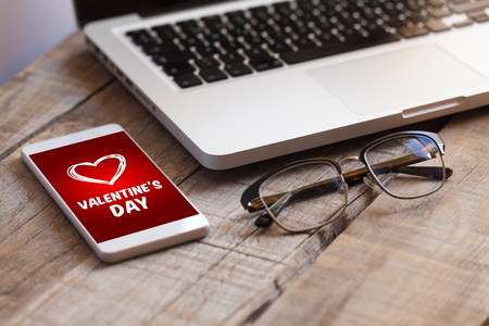 Mobile phone with Valentine's day card in the screen, with a laptop and eye glasses over a wooden table.の写真素材