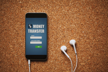 Mobile phone with money transfer app in the screen, white earphones, and copy space, on a cork panel.の写真素材