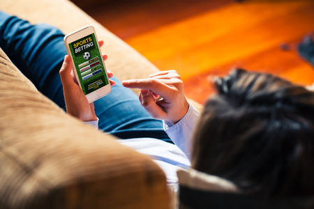 Woman holding a mobile phone to visit a sports betting website while lies down at home.の写真素材