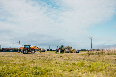Red tractor loading a trailer with straw bales during harvesting timeの写真素材