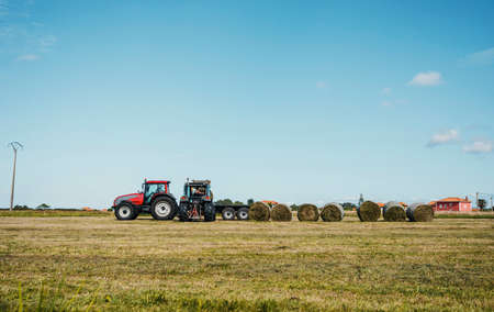 A tractor loading a trailer with straw bales during harvesting timeの写真素材