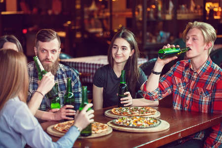 Friends having a drinks in a bar, They are sitting at a wooden table with beers and pizza. Focus on a gorgeous girl touching her bottle.の写真素材